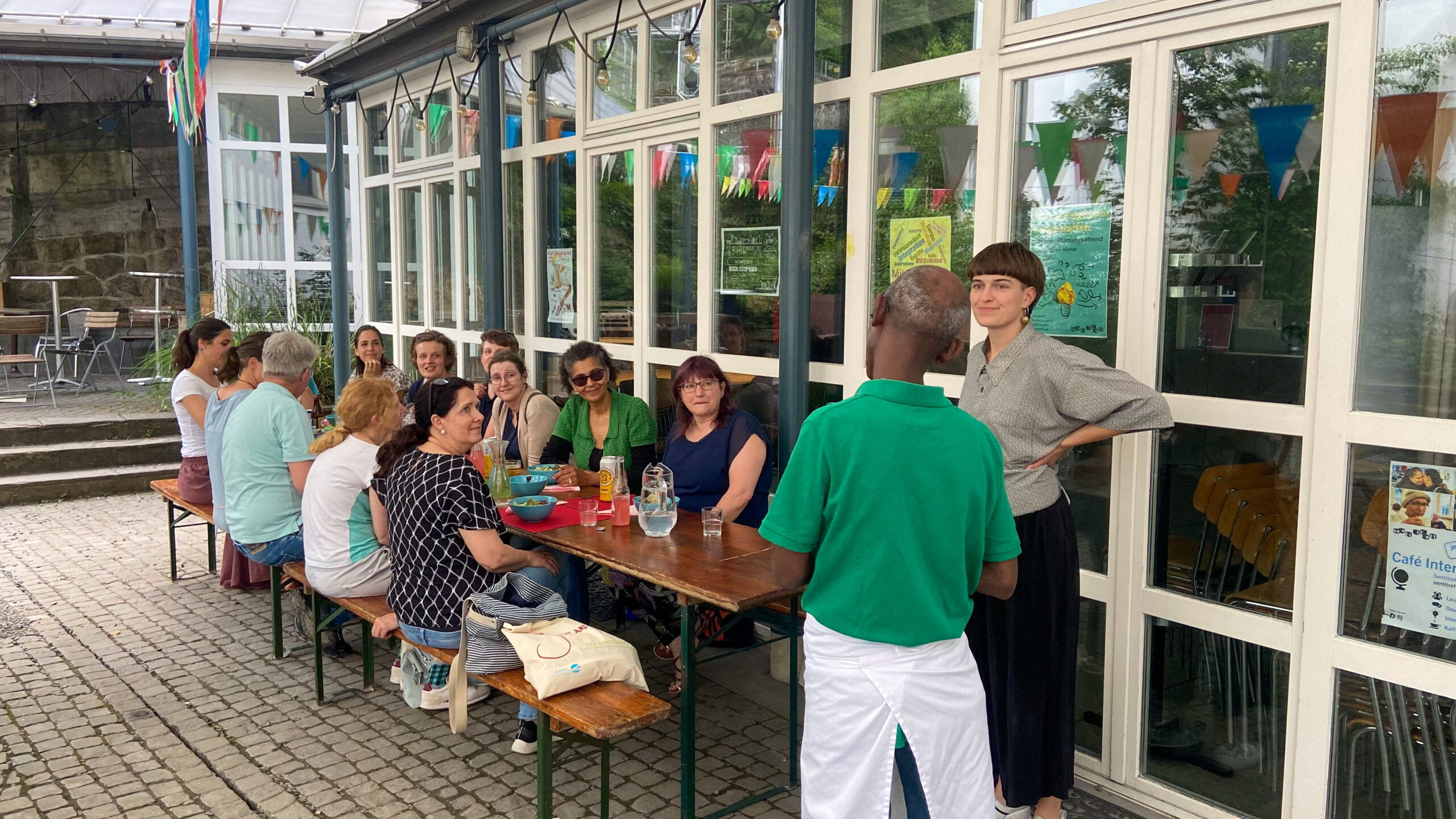 Gruppe von Menschen an einem Tisch vor einem Gebäude, daneben zwei Personen in grünen Hemden